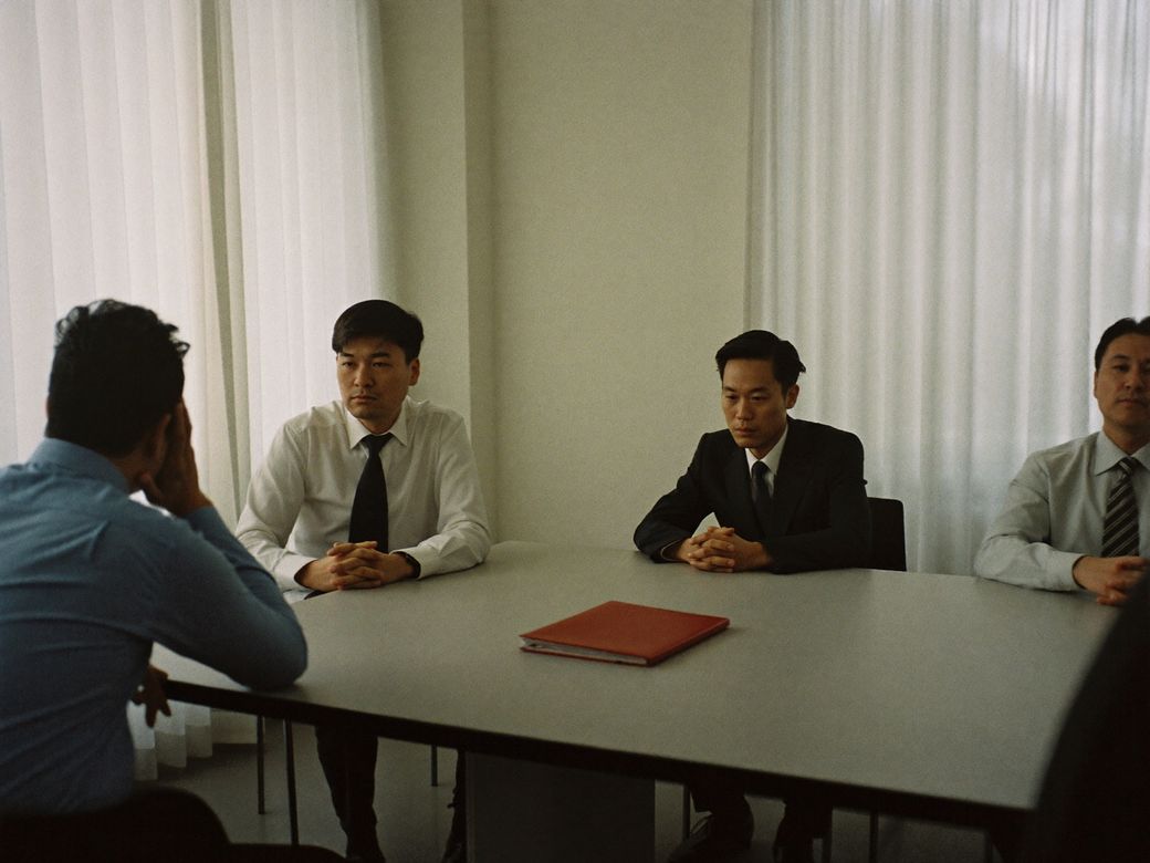 Junta de negocios donde el lado japonés escucha sin expresión y una carpeta roja descansa cerrada sobre la mesa
